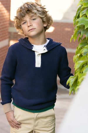 A young boy poses outdoors in a navy organic cotton henley sweater hoodie, featuring a buttoned placket and a cozy hood. The setting includes brick walls and greenery.