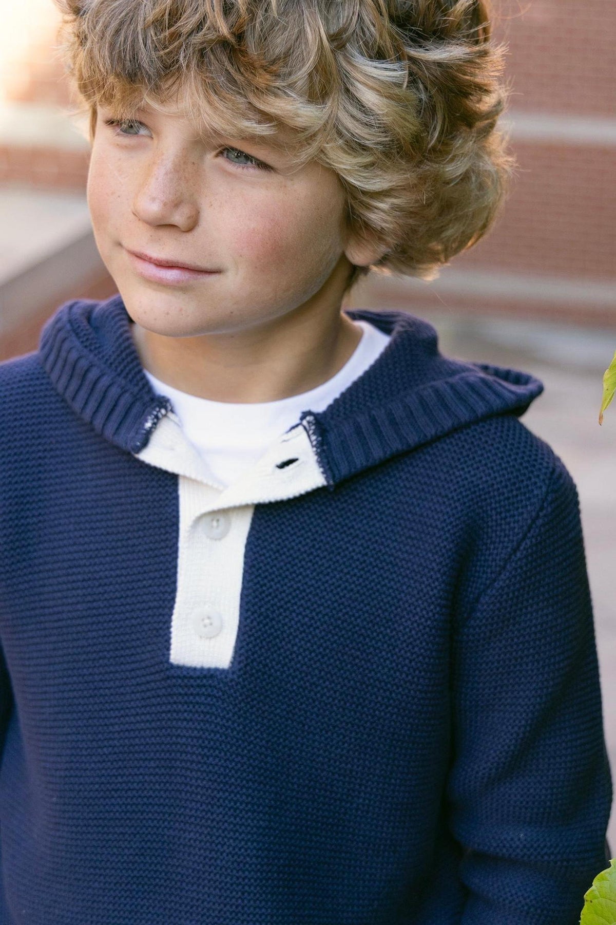 A boy with curly hair wears a navy organic cotton henley sweater hoodie with a white button placket. He stands against a brick wall, surrounded by green foliage.