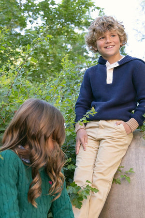 A boy wearing a navy organic cotton Henley sweater hoodie smiles while sitting on a stone amidst greenery. A girl in a green sweater is partially visible, looking down.