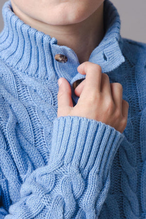 A child is wearing a medium blue organic cotton mock neck cable sweater featuring a textured knit design and ribbed trim.