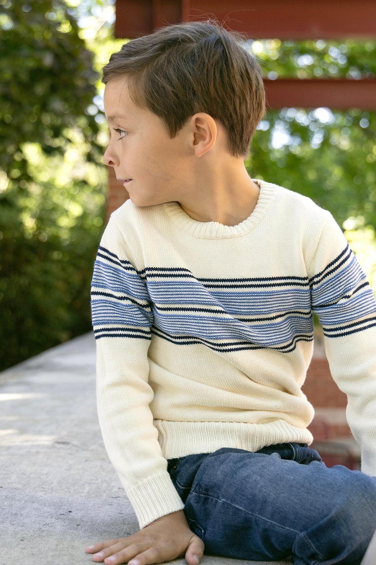 A young boy sits on a stone ledge, wearing an ivory crewneck sweater with blue stripes. The sweater is made from soft organic cotton, perfect for casual, comfortable wear.
