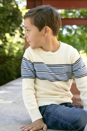 A young boy sits on a stone ledge, wearing an ivory crewneck sweater with blue stripes. The sweater is made from soft organic cotton, perfect for casual, comfortable wear.