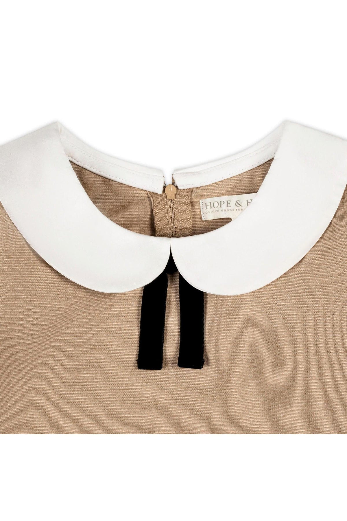 Close-up of a camel-colored dress featuring a white Peter Pan collar and a velvet bow detail, crafted from organic cotton. The label reads HOPE & J.