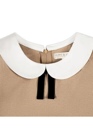 Close-up of a camel-colored dress featuring a white Peter Pan collar and a velvet bow detail, crafted from organic cotton. The label reads HOPE & J.