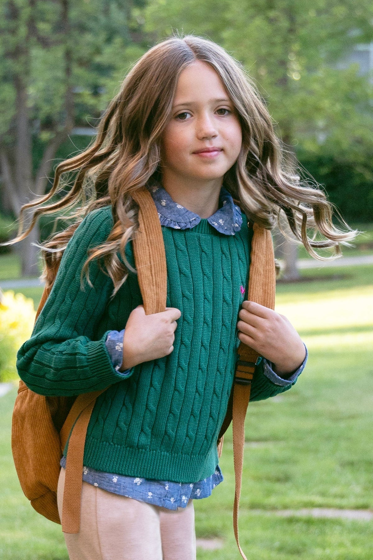 A young girl with wavy hair wears a green cable-knit sweater over a chambray shirt, carrying a brown backpack in a sunny garden setting, showcasing a casual, stylish look.