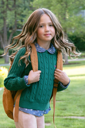A young girl with wavy hair wears a green cable-knit sweater over a chambray shirt, carrying a brown backpack in a sunny garden setting, showcasing a casual, stylish look.