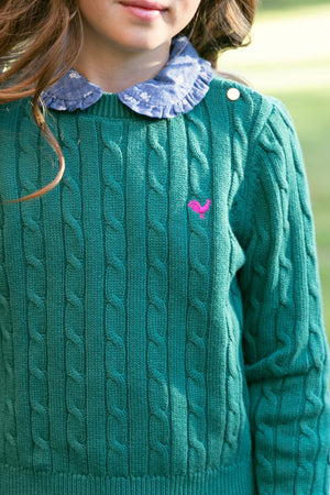 A young girl is wearing a green cable-knit sweater featuring a blue chambray Peter Pan collar. The outfit is complemented by a playful pink logo on the sweater.
