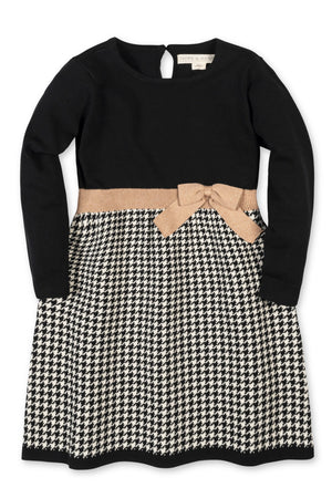 Black long-sleeve skater dress featuring a solid bodice and a houndstooth-patterned skirt. The dress is made of organic cotton and includes a decorative bow at the waist.