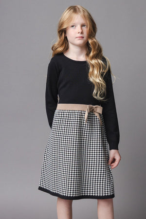 A girl stands against a gray backdrop wearing a long-sleeve black dress with a houndstooth skirt and a decorative bow at the waist. The dress is made from organic cotton.