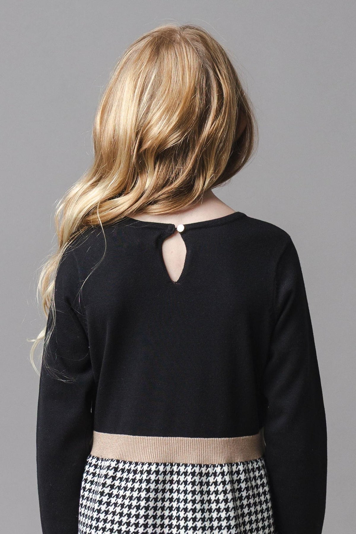 A young girl with long, wavy blonde hair is seen from the back, wearing a black sweater dress featuring a decorative keyhole closure at the neckline and a houndstooth skirt.