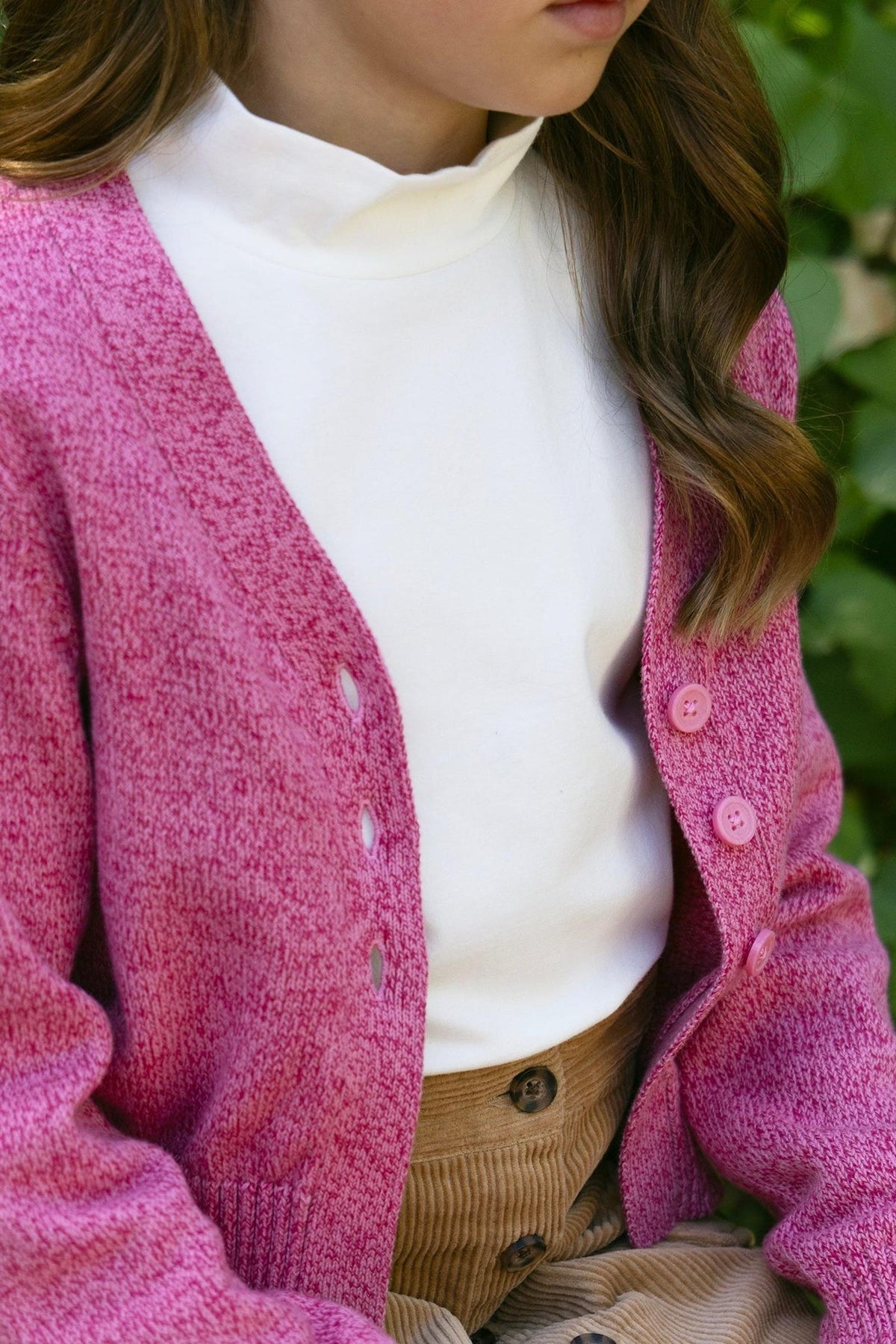 A young girl sits on a wooden bench, wearing a vibrant pink organic cotton V-neck cardigan over a white turtleneck, complemented by a pair of brown corduroy pants amidst lush.