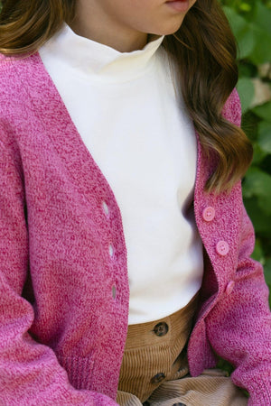 A young girl sits on a wooden bench, wearing a vibrant pink organic cotton V-neck cardigan over a white turtleneck, complemented by a pair of brown corduroy pants amidst lush.