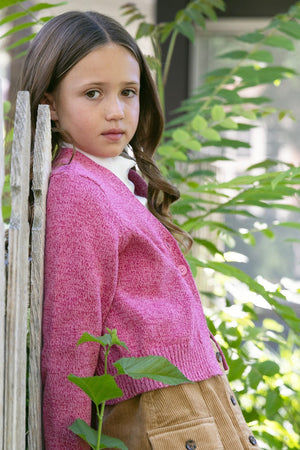 A young girl leans against a wooden fence, wearing a vibrant pink cardigan. The setting features lush greenery, highlighting the soft, organic cotton material of the garment.