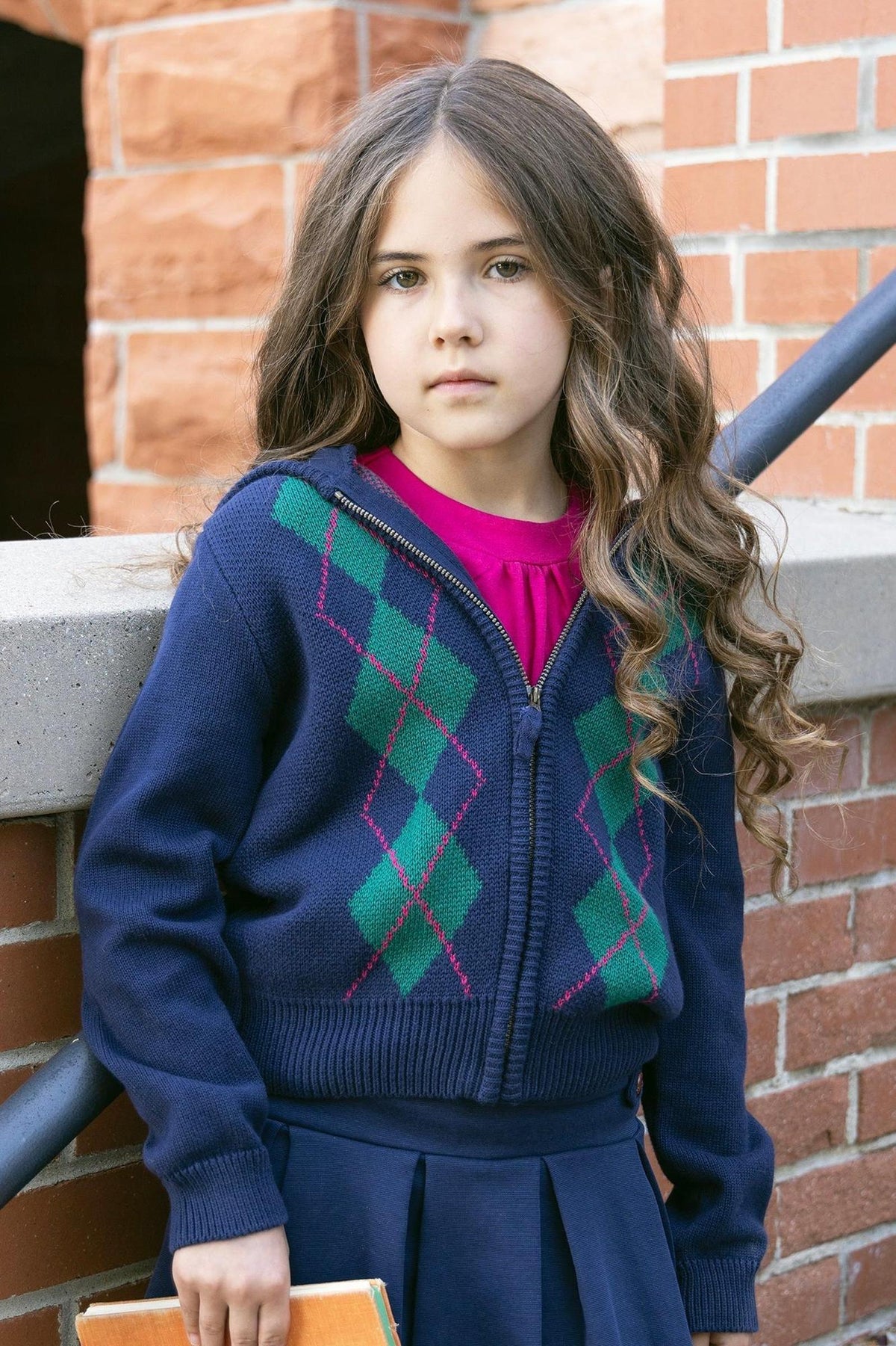 A young girl stands against a brick wall, wearing a navy zip-up hoodie with a green and purple argyle pattern.