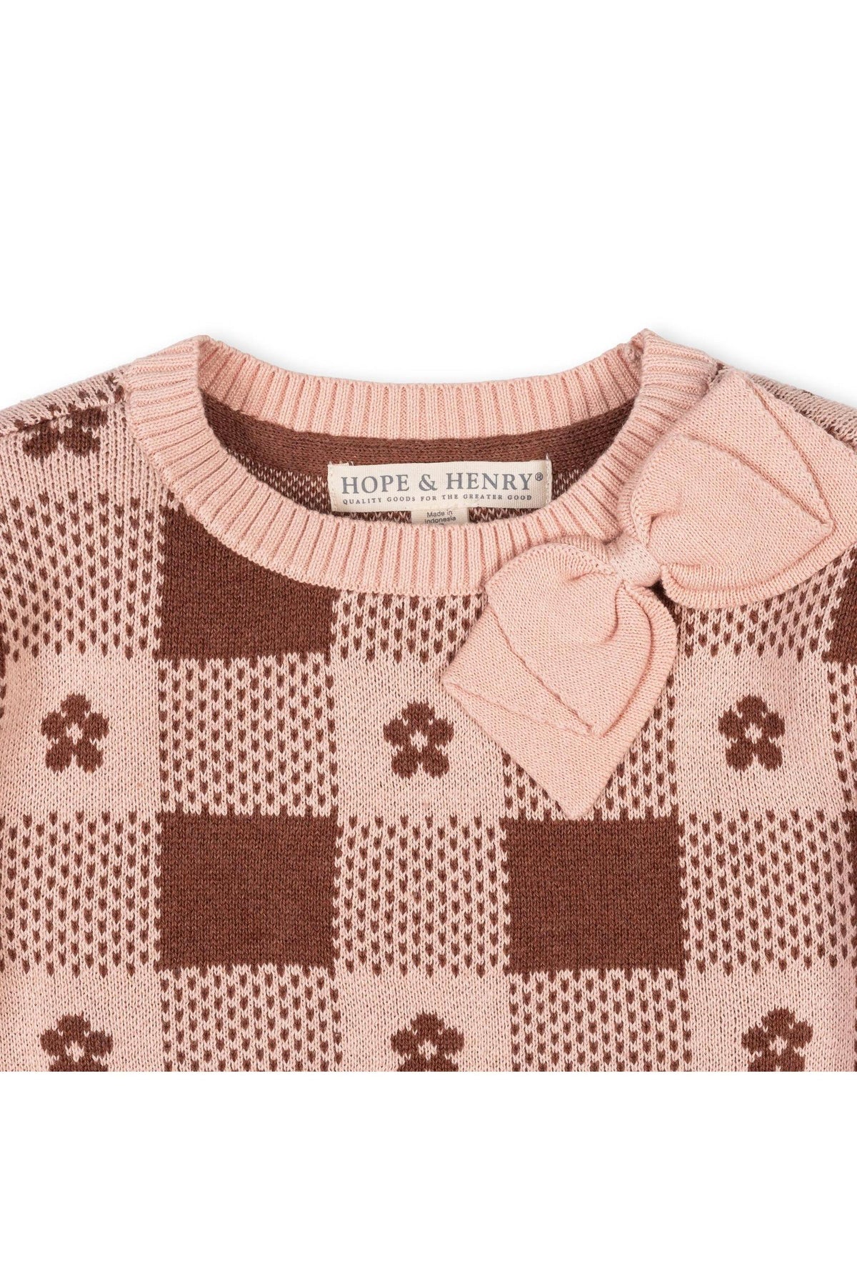 A close-up of a knitted sweater featuring a pink and brown checkered pattern, adorned with a bow detail. It is labeled with HOPE & HENRY, made from soft organic cotton.