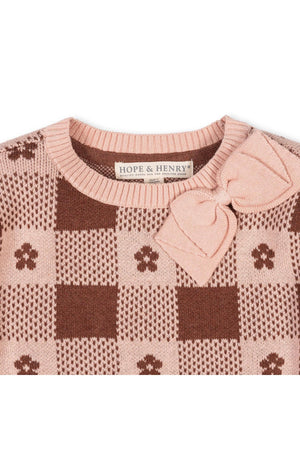 A close-up of a knitted sweater featuring a pink and brown checkered pattern, adorned with a bow detail. It is labeled with HOPE & HENRY, made from soft organic cotton.