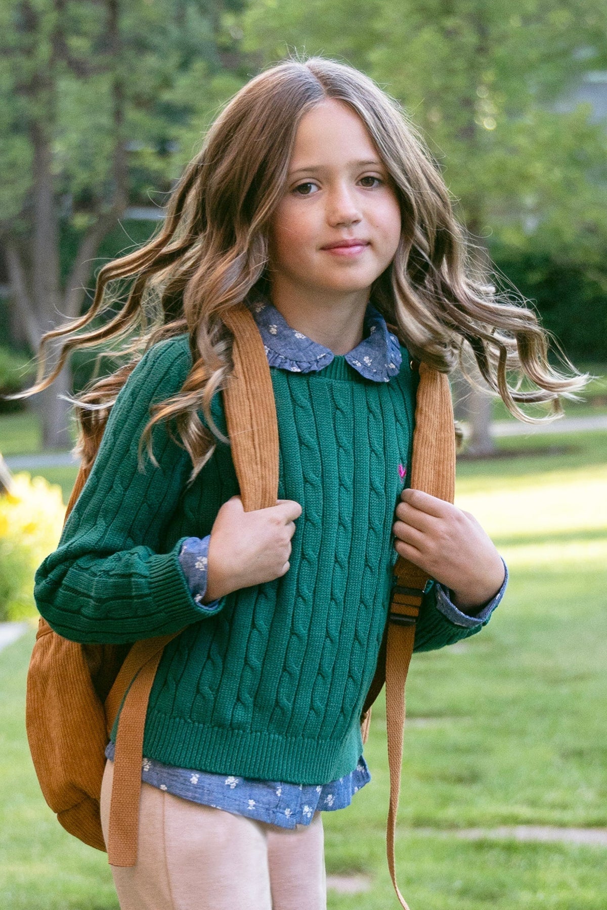 A young girl with wavy hair wears a green cable knit sweater and carries a brown backpack, standing in a lush garden with vibrant greenery and sunlight in the background.
