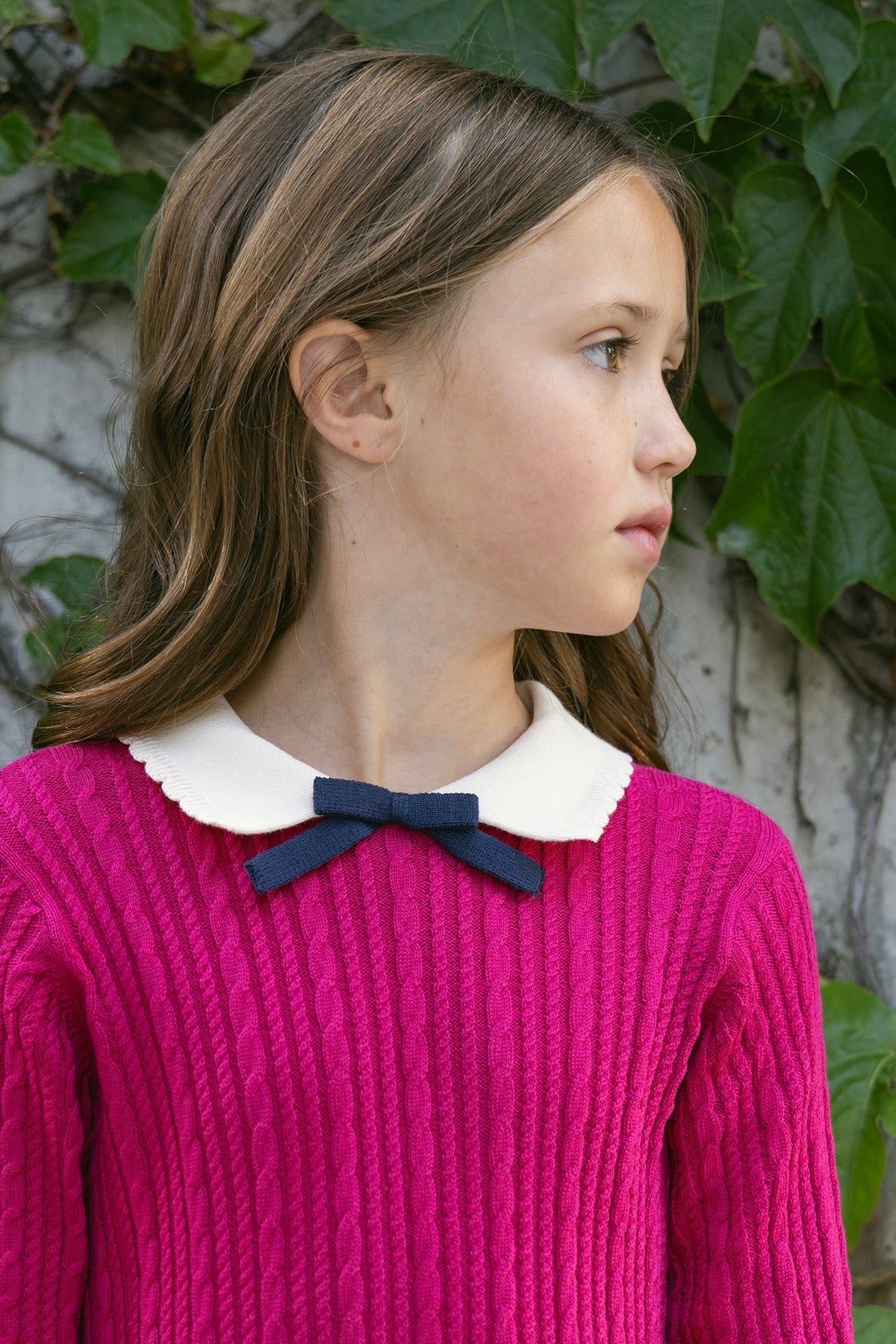 A young girl stands against a backdrop of green ivy, wearing a vibrant pink cable knit sweater dress with a white scalloped collar and a navy bow.