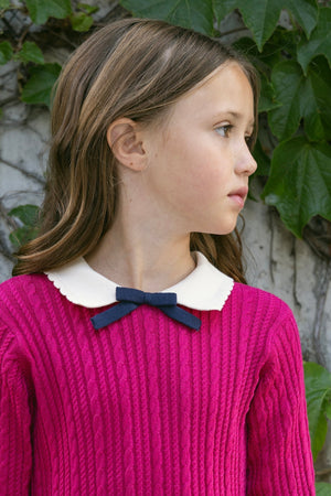 A young girl stands against a backdrop of green ivy, wearing a vibrant pink cable knit sweater dress with a white scalloped collar and a navy bow.