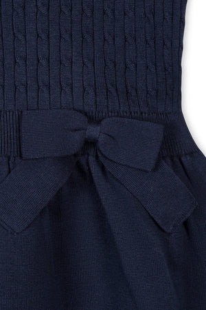 Close-up image of a navy blue cable knit sweater dress featuring a textured bodice and a bow detail at the waist, made from 100% GOTS Certified organic cotton for comfort.