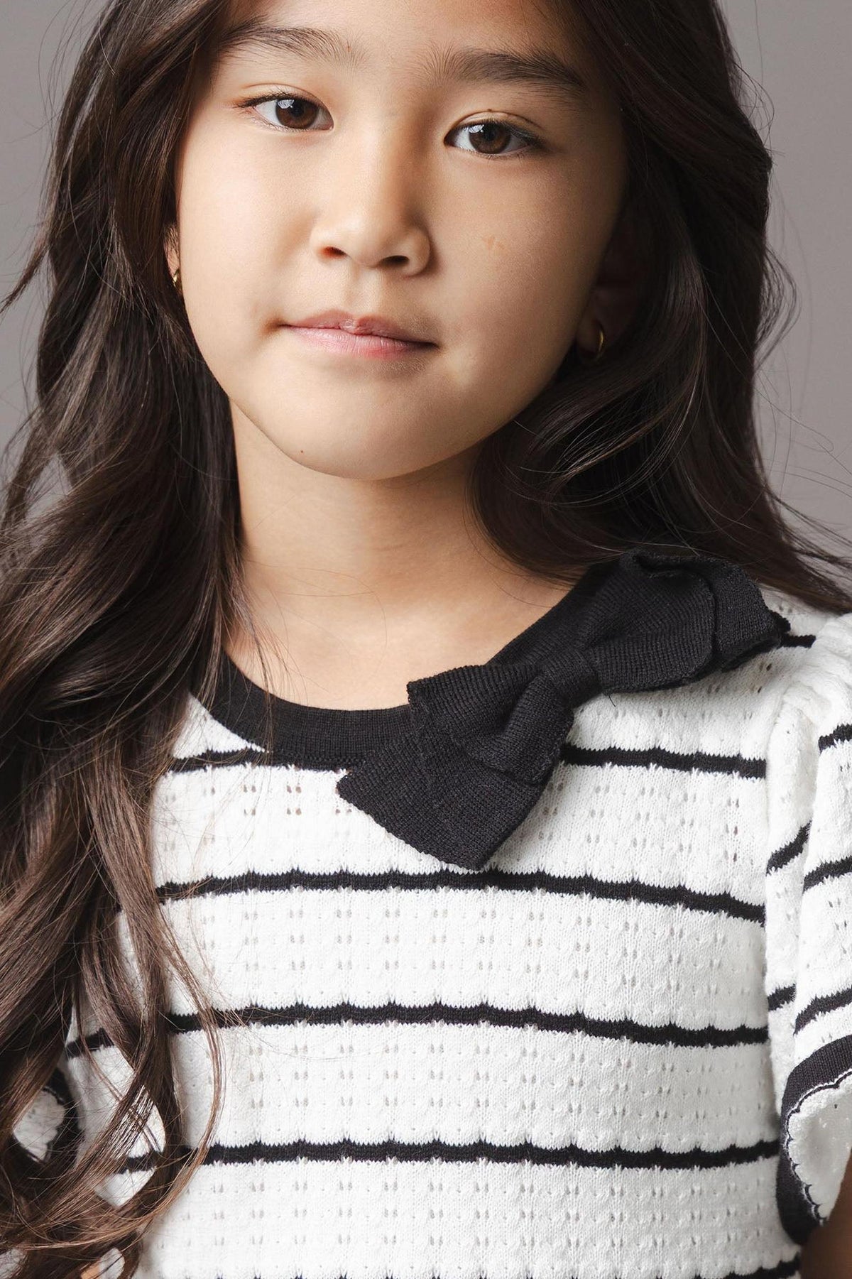 A young girl models a black and white striped sweater dress with short sleeves and a bow detail at the neckline.