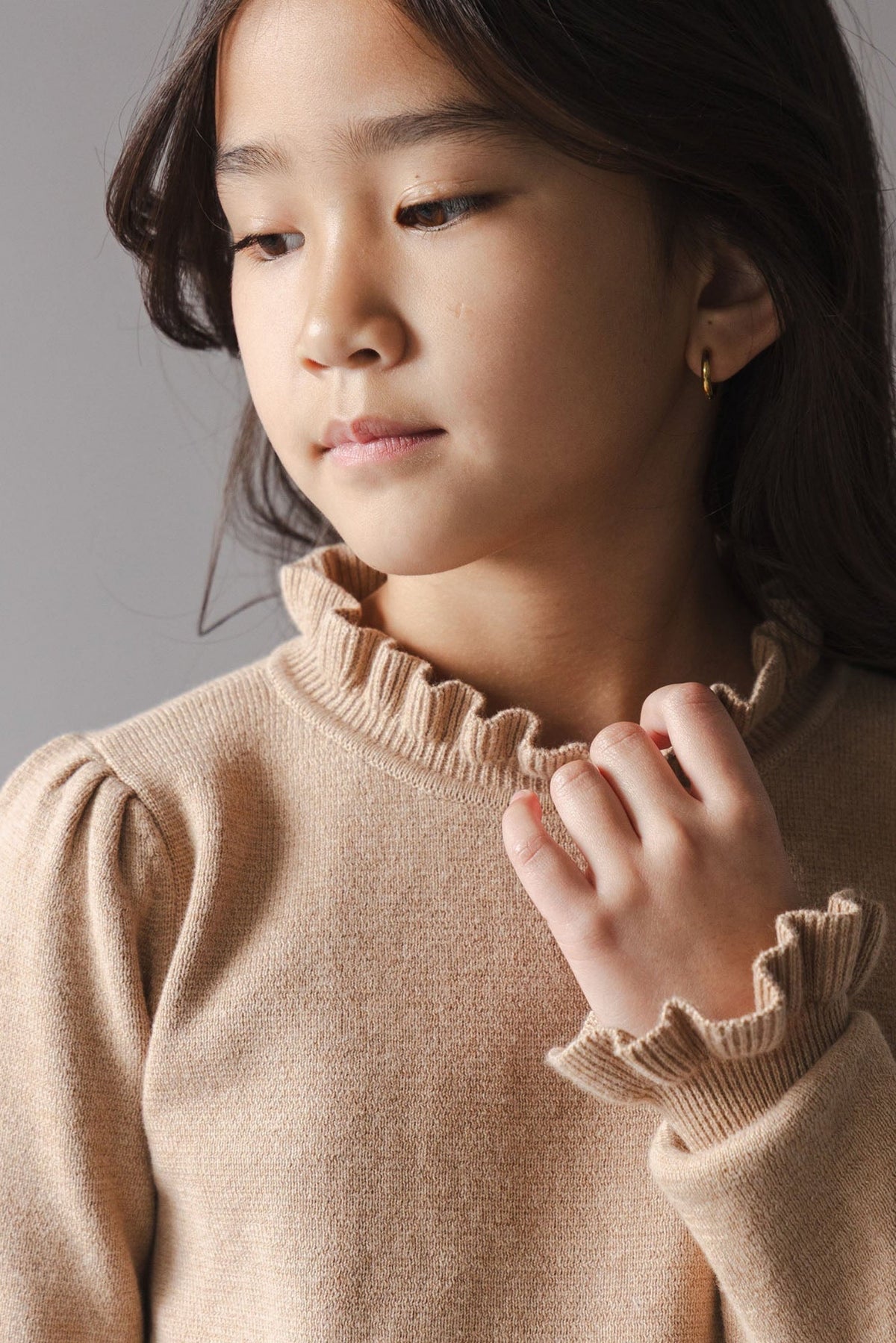A young girl models a cozy camel-colored sweater dress made from organic cotton. The dress features a ruffled mock neck and cuffs, showcasing a blend of comfort and style.