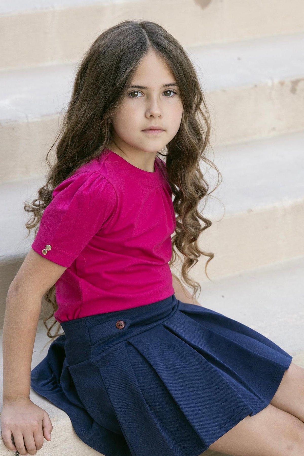 A young girl with long, wavy hair sits on steps wearing a bright pink shirt and a classic navy pleated skort.