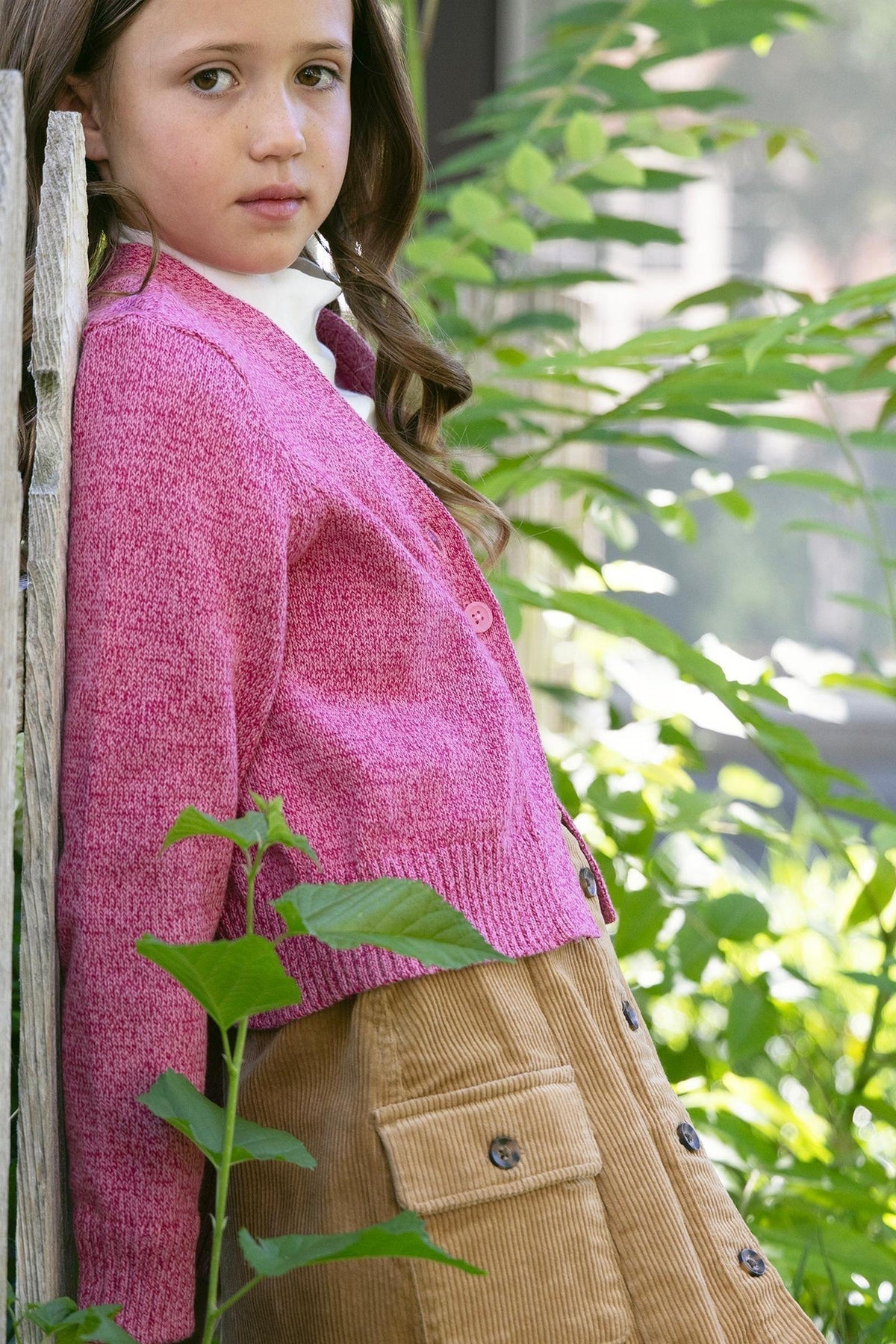 A girl with long brown hair leans against a wooden fence, wearing a pink cardigan and a brown organic cotton corduroy skirt.