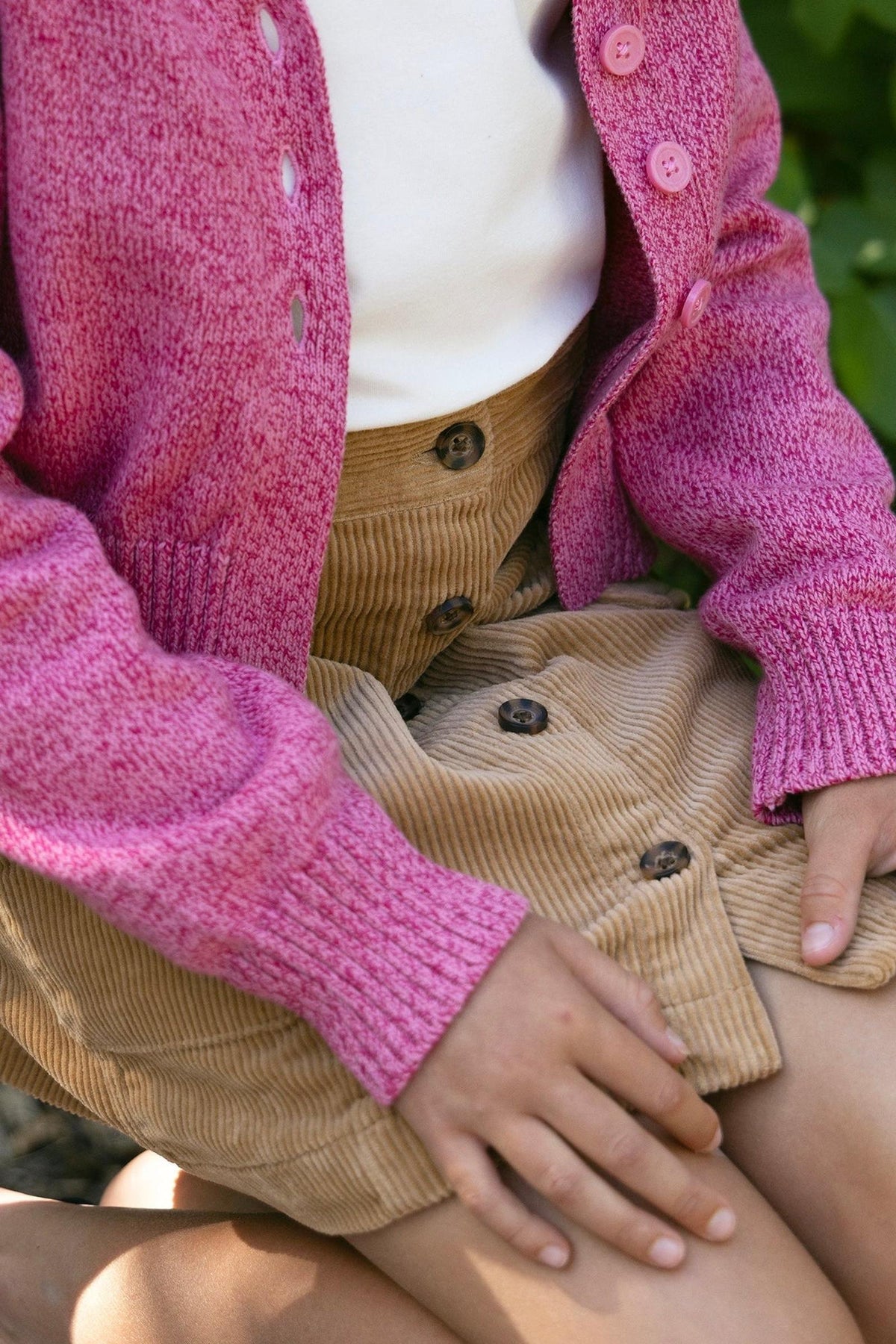 A child sits on a wooden bench, wearing a pink cardigan over a cream top and a brown organic cotton corduroy button-front skirt.