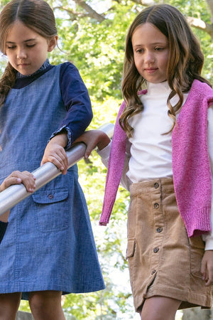 Two young girls play outdoors, one wearing a denim dress and the other in a brown corduroy button-front skirt paired with a pink sweater.