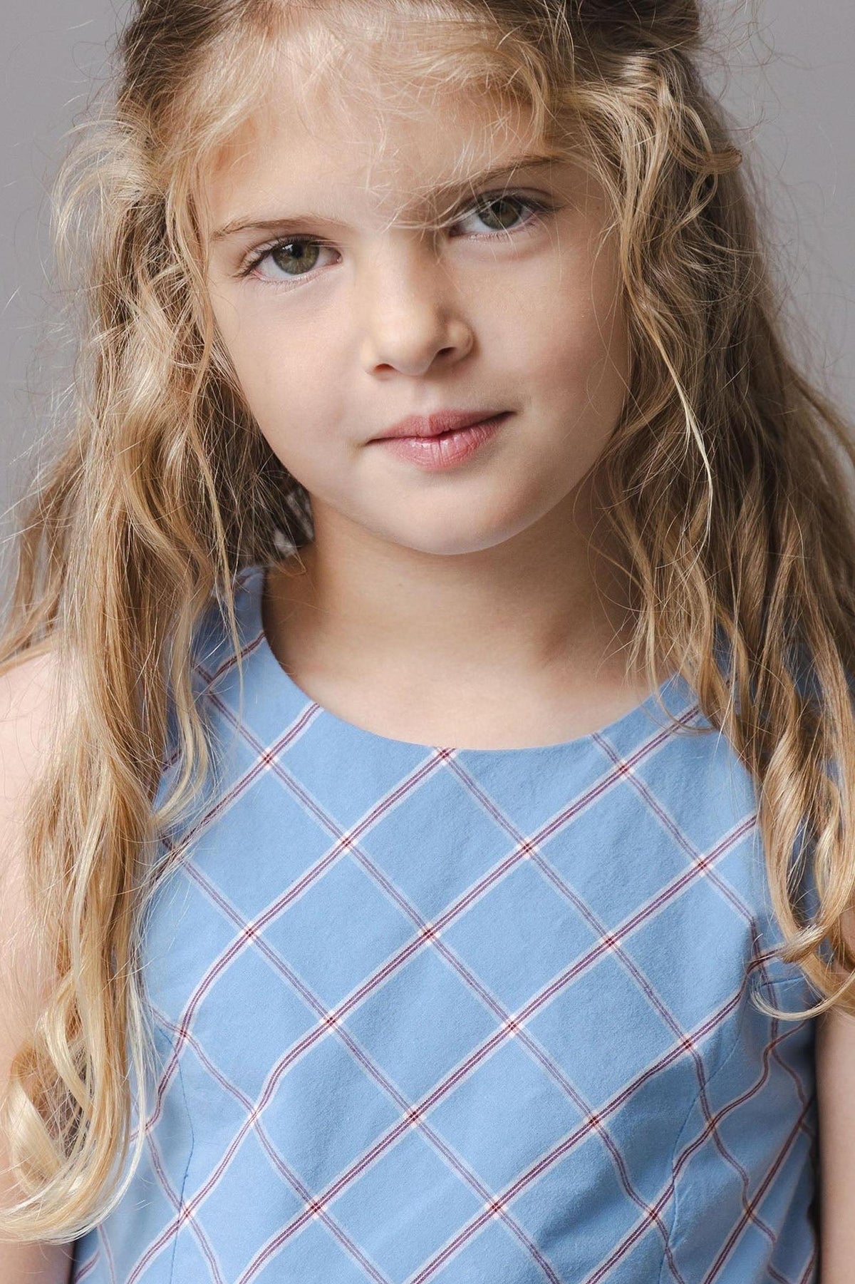 A young girl with long, wavy hair is wearing a sleeveless blue dress featuring a plaid pattern. The dress has a soft, organic cotton fabric, perfect for comfort and movement.