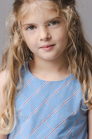 A young girl with long, wavy hair is wearing a sleeveless blue dress featuring a plaid pattern. The dress has a soft, organic cotton fabric, perfect for comfort and movement.