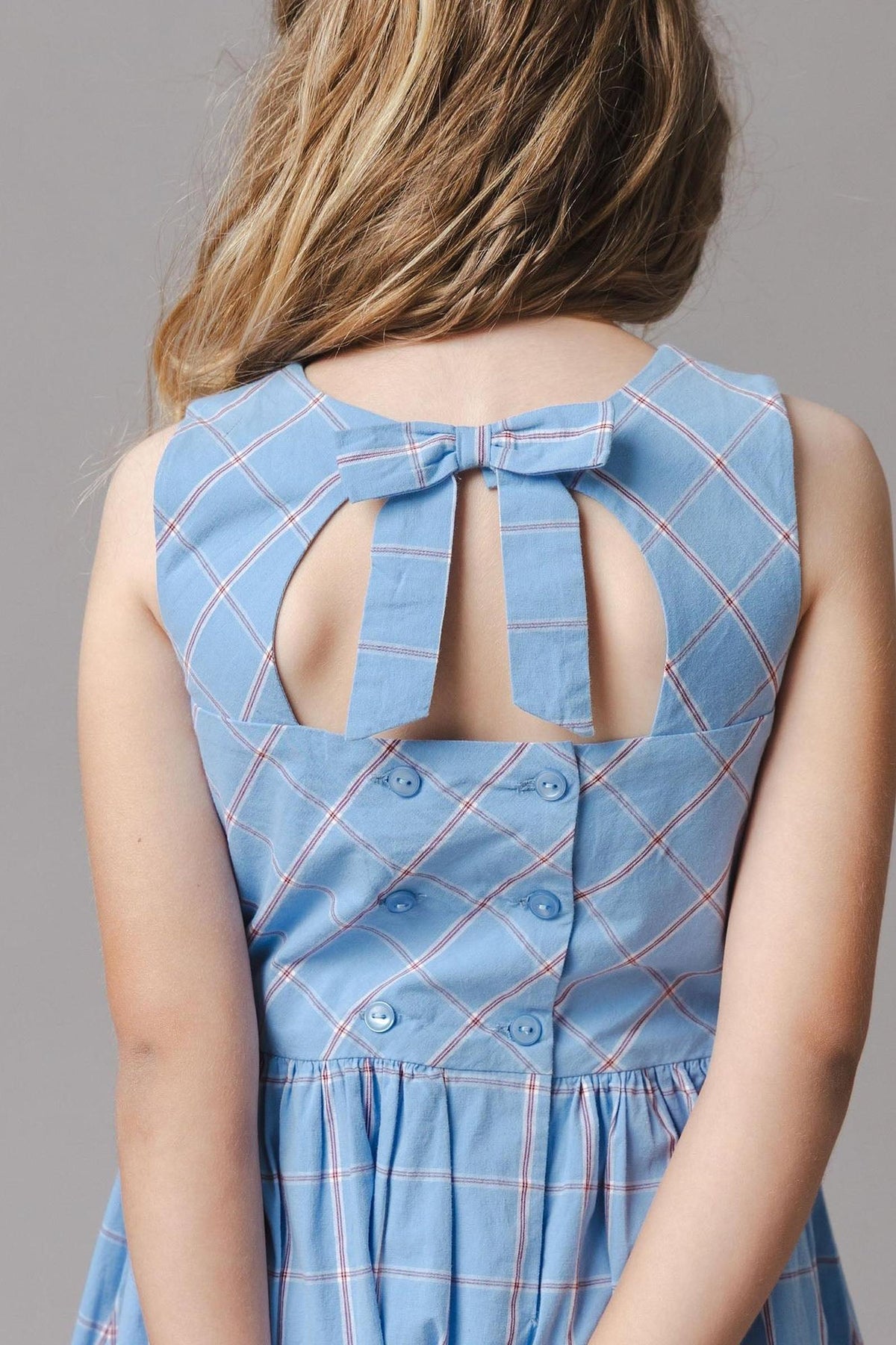 A young girl shows the back of a sleeveless blue plaid dress featuring a bow and button closure. The dress is made from organic cotton, designed for comfort and style.