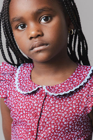 A young girl models a wine-colored dress featuring a Peter Pan collar and flutter sleeves, adorned with a delicate floral pattern. The dress is styled with a button front closure.