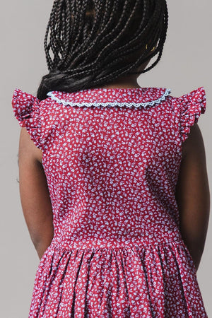 A young girl is seen from behind wearing a wine-colored linen dress adorned with a delicate floral pattern. The dress features a Peter Pan collar and sweet flutter sleeves.