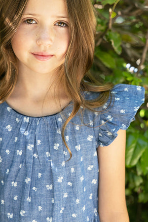 A young girl stands amidst lush greenery, wearing a blue chambray dress with ruffle sleeves and white floral patterns. The dress is made from soft, breathable organic cotton.