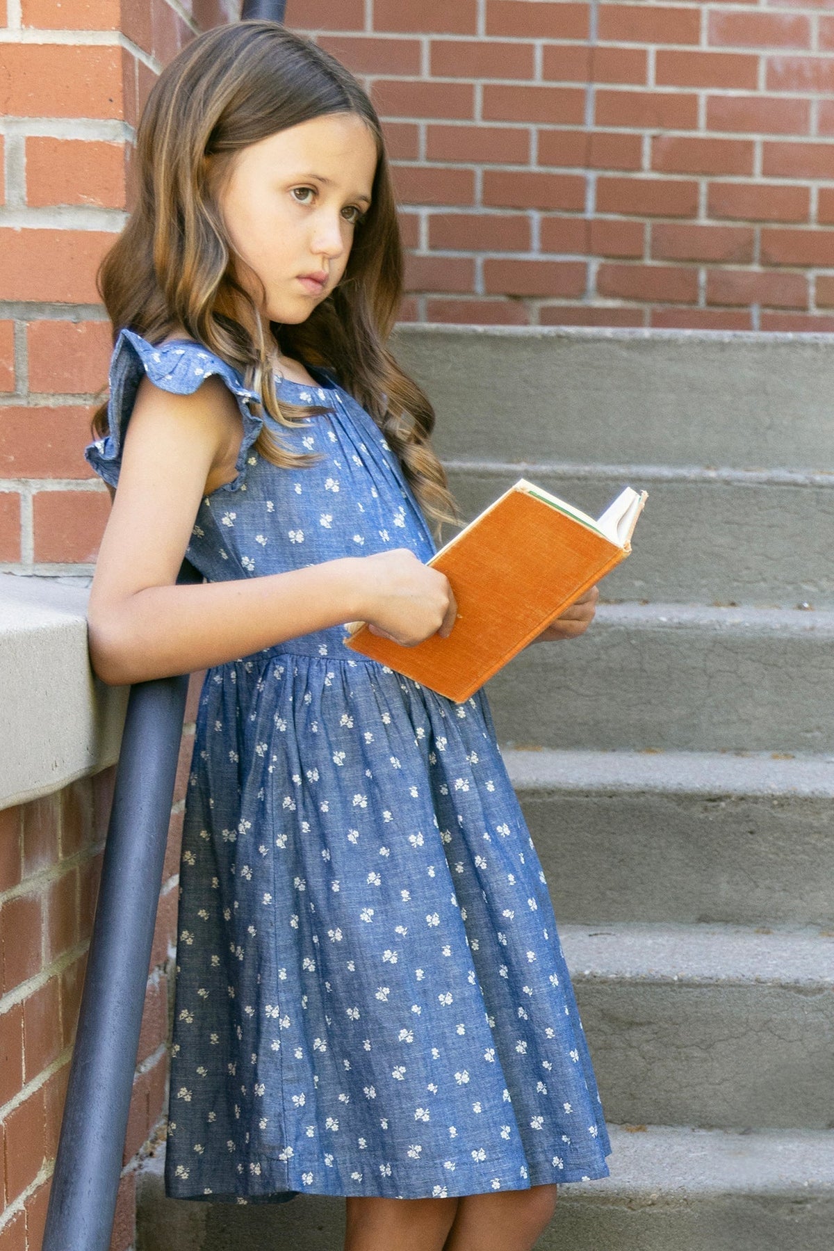 A young girl stands on stone steps, wearing a blue chambray dress with ruffle sleeves and white floral patterns, holding an orange book. The setting features a brick wall backdrop.