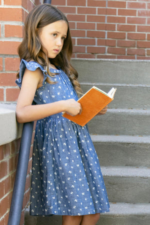 A young girl stands on stone steps, wearing a blue chambray dress with ruffle sleeves and white floral patterns, holding an orange book. The setting features a brick wall backdrop.