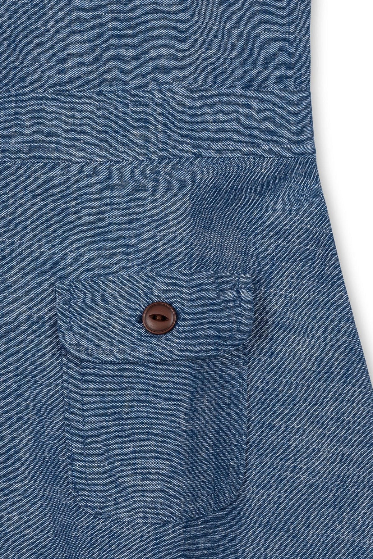Close-up of a chambray fabric featuring a small chest pocket with a brown button. The soft, organic cotton material showcases a light blue hue, emphasizing a classic and timeless style.