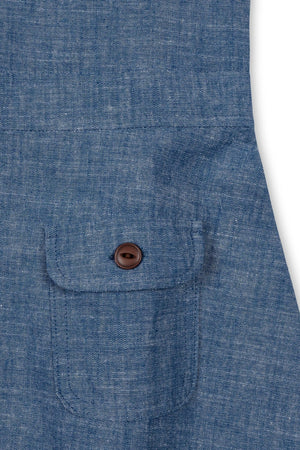 Close-up of a chambray fabric featuring a small chest pocket with a brown button. The soft, organic cotton material showcases a light blue hue, emphasizing a classic and timeless style.