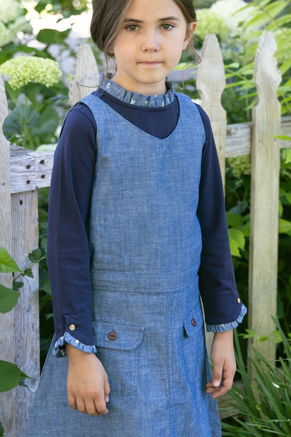 A young girl stands outdoors, wearing a chambray jumper with a V-neck over a navy long-sleeve shirt. The setting features greenery and a wooden fence, highlighting a natural, playful atmosphere.