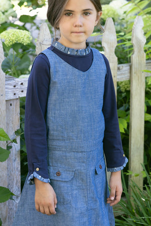A young girl stands outdoors, wearing a chambray jumper with a V-neck over a navy long-sleeve shirt. The setting features greenery and a wooden fence, highlighting a natural, playful atmosphere.