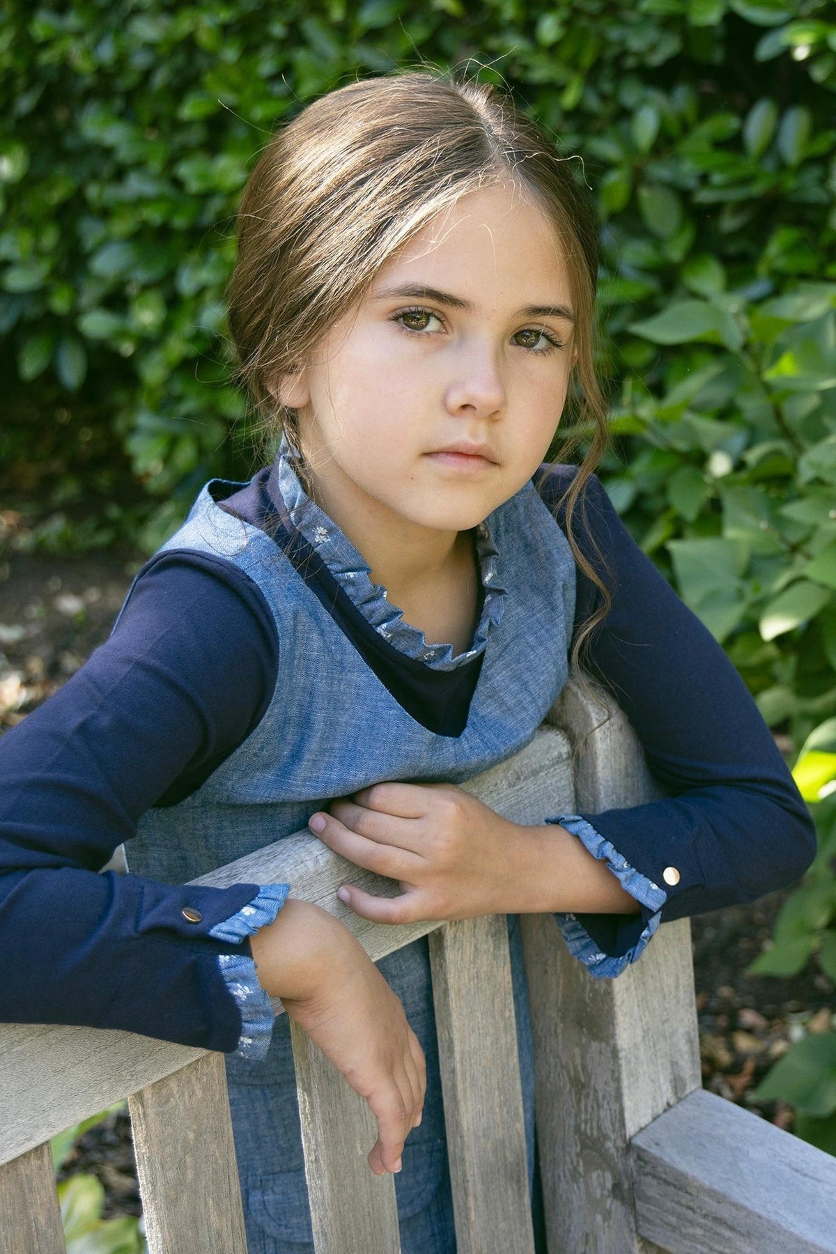 A young girl with long hair sits on a wooden bench, wearing a chambray jumper and a navy long-sleeve shirt.