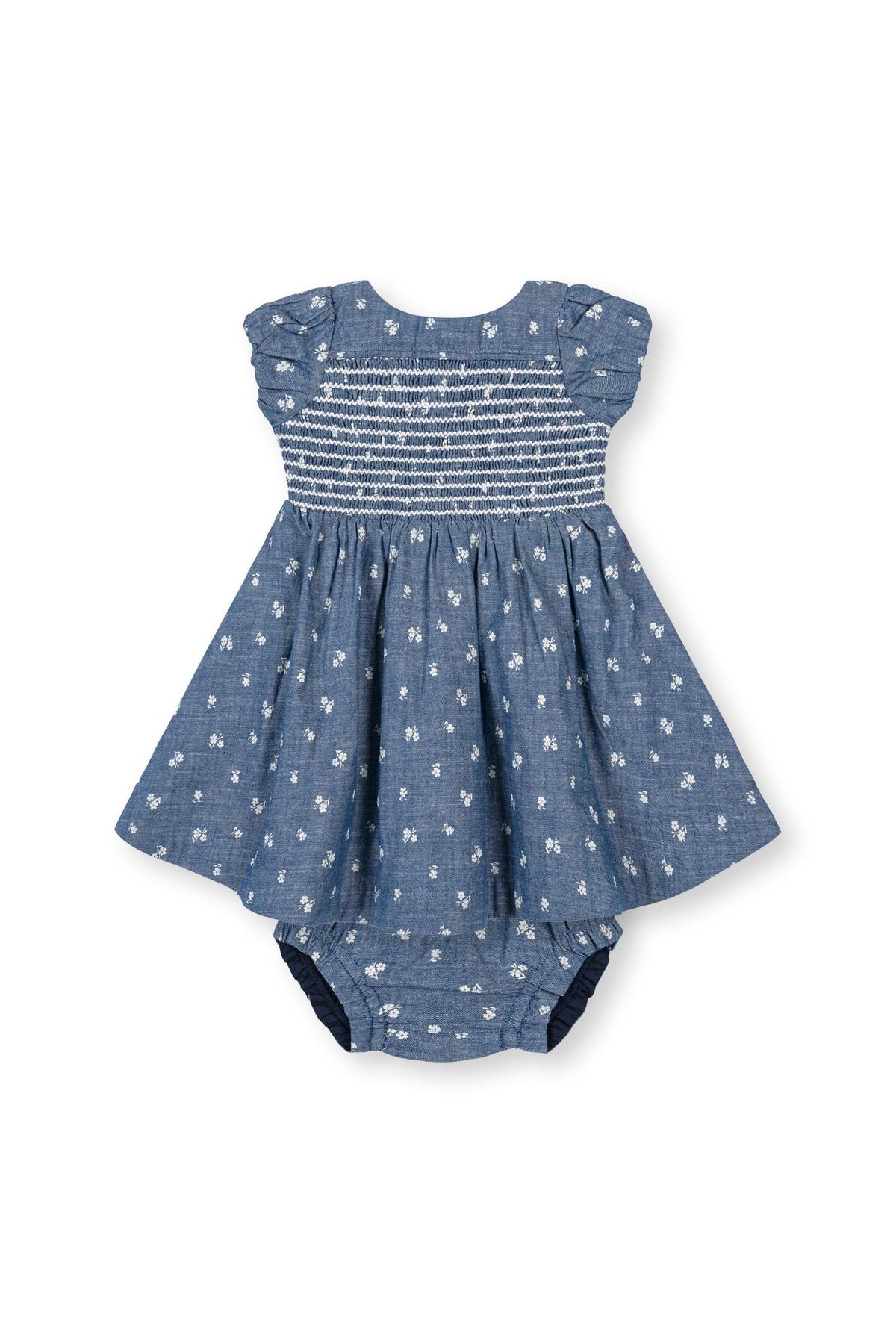 A blue chambray baby dress features smocked bodice and puff sleeves, decorated with delicate white floral embroidery. Made from 100% organic cotton, it offers a soft, breathable fabric for comfort.