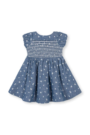 A soft blue chambray baby dress features a smocked bodice and delicate floral embroidery. Crafted from 100% organic cotton, this dress is perfect for special occasions and everyday wear.