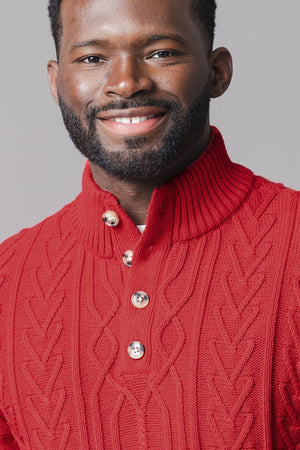 A smiling man wears a red mock neck cable sweater made from soft organic cotton, featuring a ribbed collar.