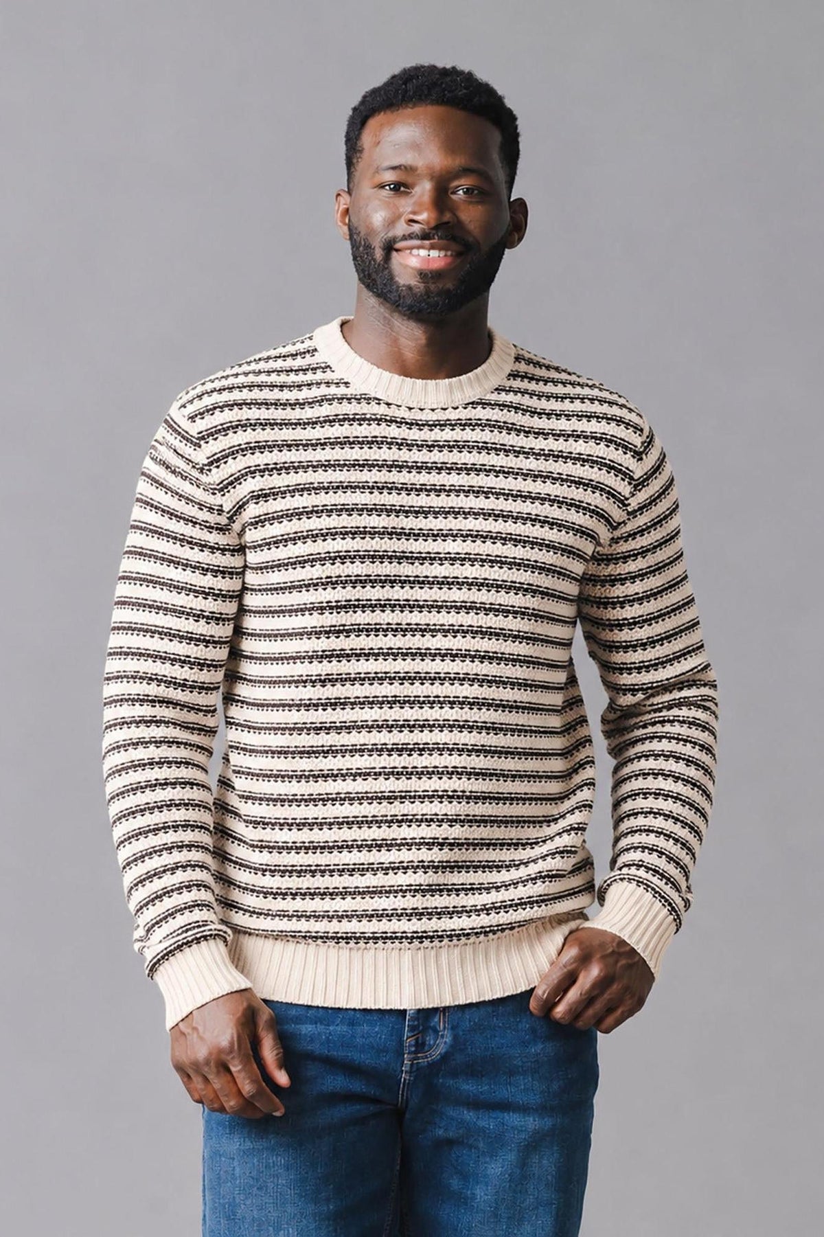 A man poses in a striped crew neck sweater featuring brown and tan organic cotton. The relaxed fit and textured knit add a stylish touch to casual wear.