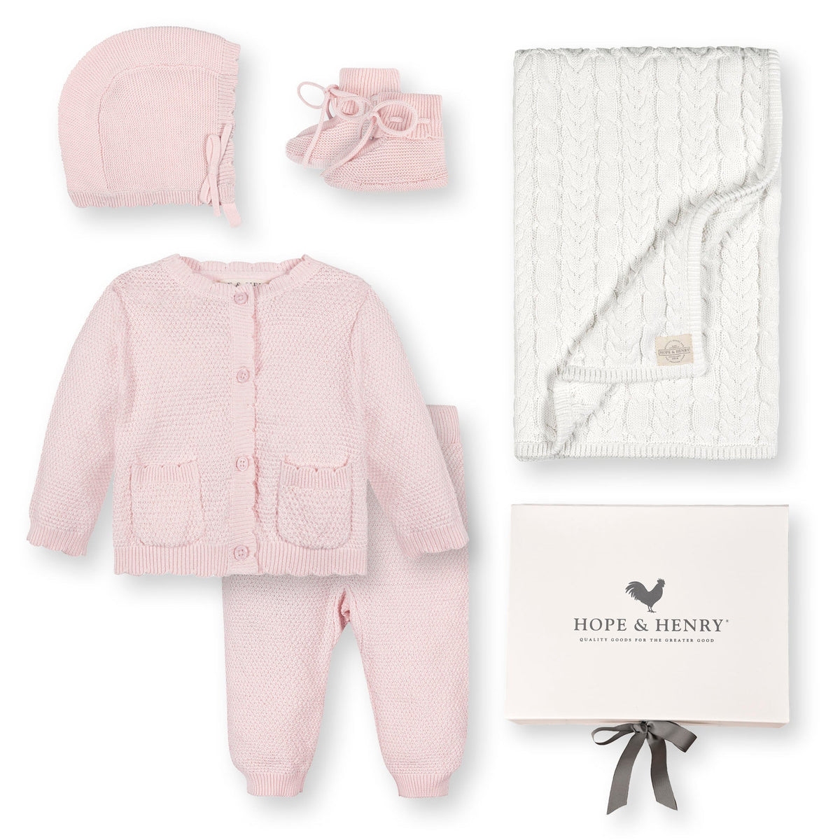 A pale pink baby sweater set by Hope & Henry includes a cardigan, bonnet, booties, and leggings, accompanied by a cream cable-knit blanket, all crafted from soft organic cotton.