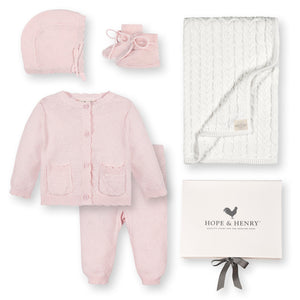A pale pink baby sweater set by Hope & Henry includes a cardigan, bonnet, booties, and leggings, accompanied by a cream cable-knit blanket, all crafted from soft organic cotton.