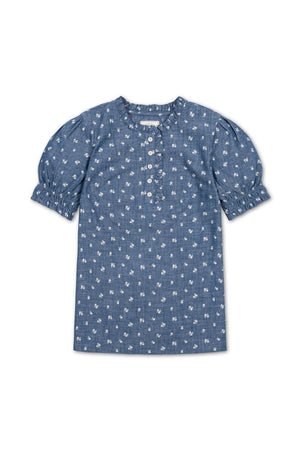 A blue chambray puff sleeve top features a delicate floral print, ruffle trim at the collar, and buttons down the front, crafted from 100% organic cotton for comfort.